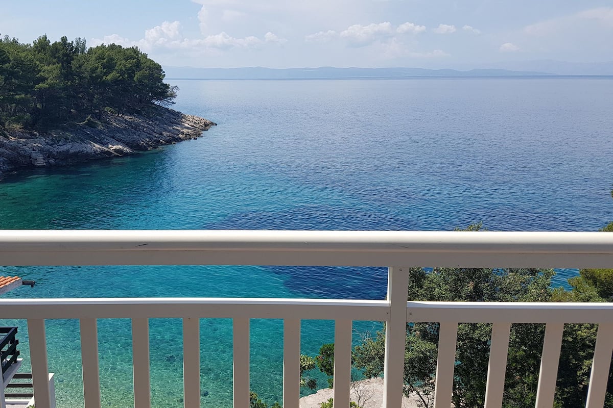 A balcony with a view of a clear blue sea and a rocky coastline with trees.