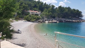 A pebble beach with clear turquoise water, a stone house in the background, and a metal railing in the foreground. - Apartments Teo i Ivana (Bogomolje)