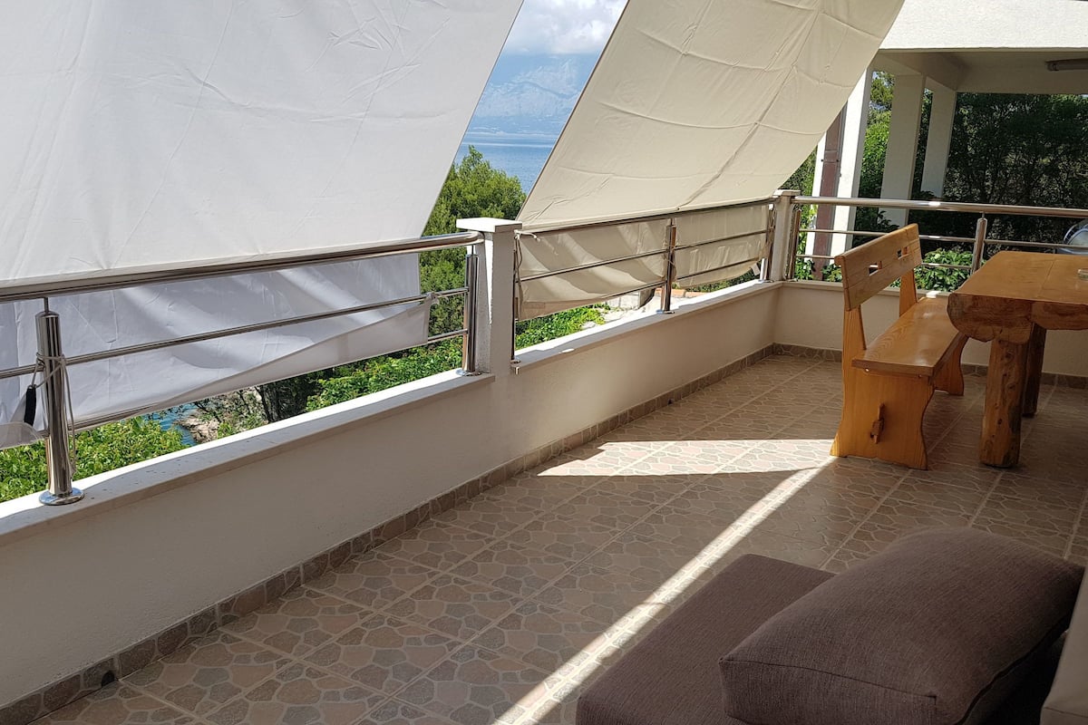 A balcony with a view of a body of water and greenery, a wooden table, and a bench.
