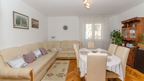 A living room with a beige sofa, a dining table with chairs, a wooden cabinet, and a patterned rug. - Apartments Kata (Supetar)