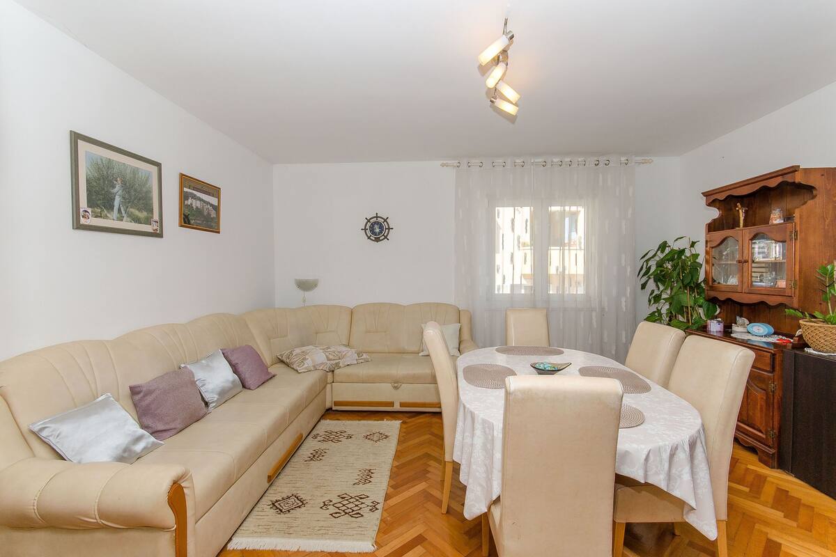 A living room with a beige sofa, a dining table with chairs, a wooden cabinet, and a patterned rug.