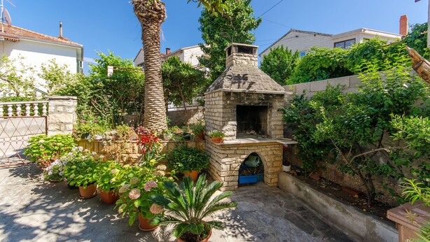 A stone-built outdoor kitchen with a chimney, surrounded by potted plants and a palm tree. - Apartments Kata (Supetar)