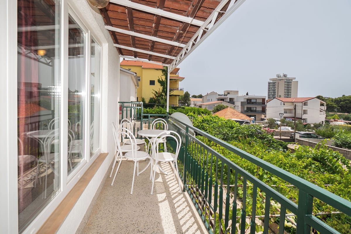 A balcony with white chairs and tables, a green railing, and a view of surrounding buildings.