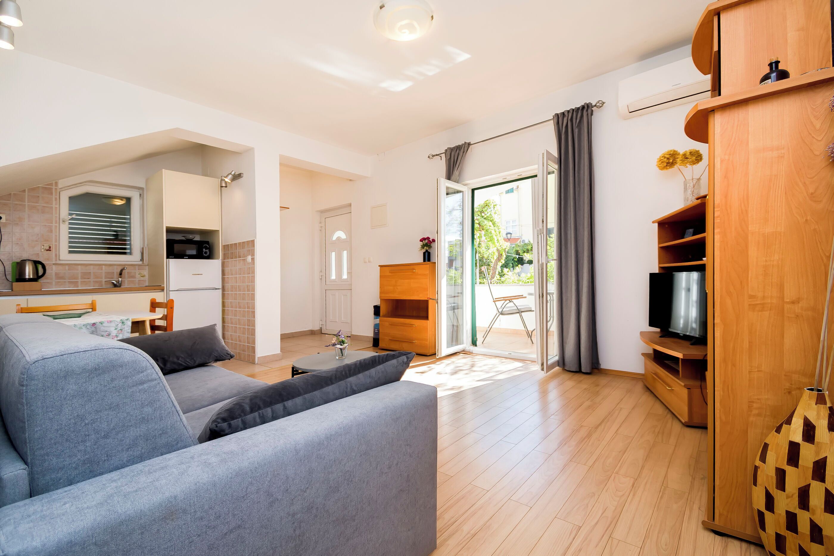 A modern living room with a grey sofa, wooden flooring, a TV stand, and a sliding glass door leading to an outdoor area.