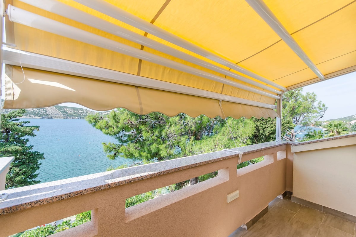A balcony with a view of a lake and trees, a yellow awning, and a tiled floor.