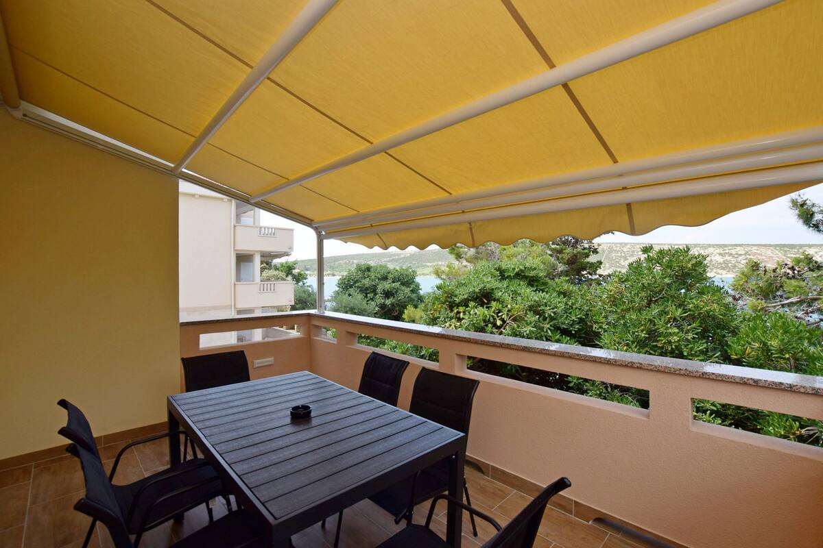 A balcony with a table and chairs, a yellow canopy, and a view of greenery and buildings.