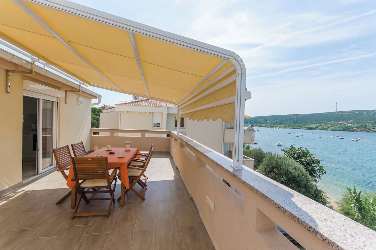 A balcony with a table and chairs, a yellow awning, and a view of boats on a body of water.