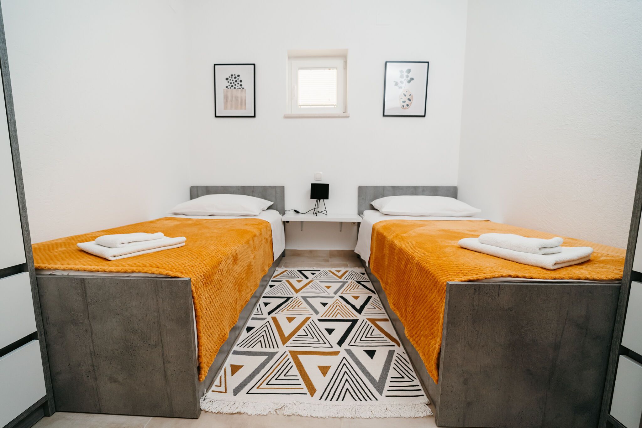 A room with two beds, each with a gray headboard and an orange blanket. There is a patterned rug on the floor, a small table with a lamp, and two framed pictures on the wall.
