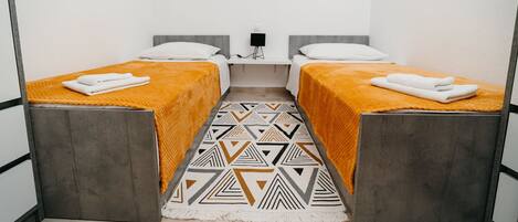 A room with two beds, each with a gray headboard and an orange blanket. There is a patterned rug on the floor, a small table with a lamp, and two framed pictures on the wall.