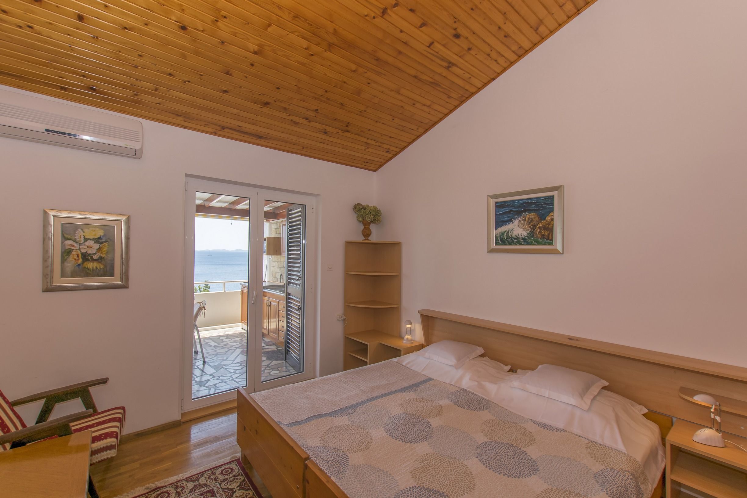 A bedroom with a wooden ceiling, a bed with a patterned bedspread, a nightstand with a lamp, a window with a view, and a door leading to a balcony.