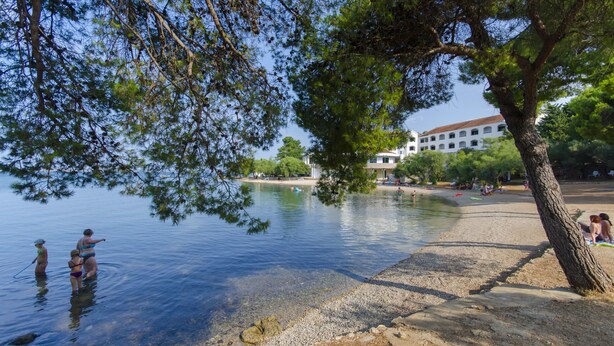 A lakeside scene with clear water, a sandy beach, and a building in the background. - Apartments Andrija (Pirovac)
