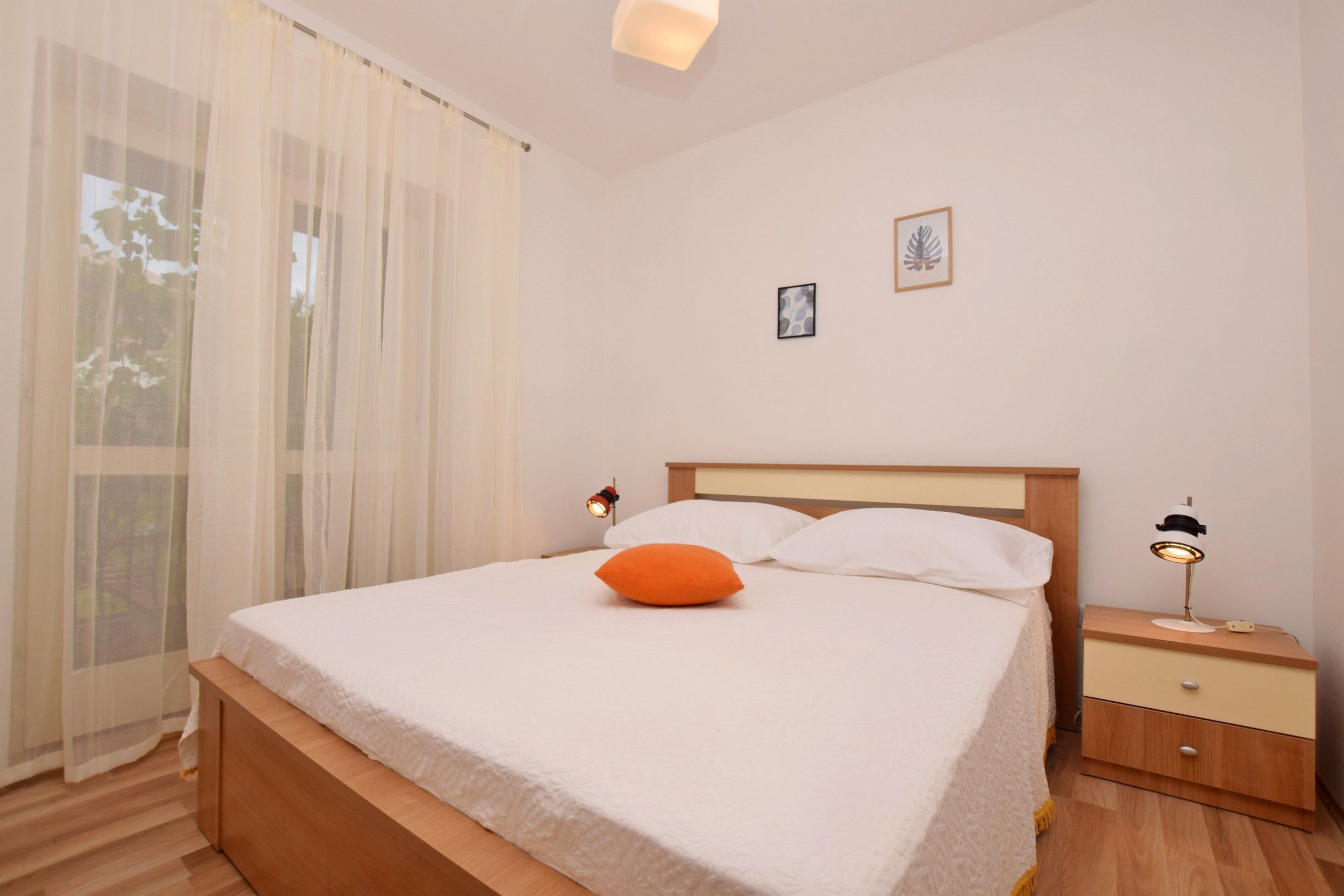 Un lit bien fait, avec une literie blanche et un oreiller orange, une table de chevet avec une lampe, et une fenêtre ornée de rideaux légers.
