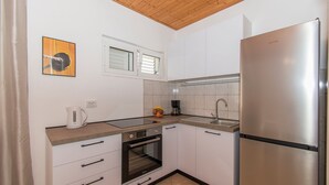 A modern kitchen with white cabinets, a stainless steel refrigerator, a built-in oven, and a window with blinds. - Apartments Andrija (Pirovac)