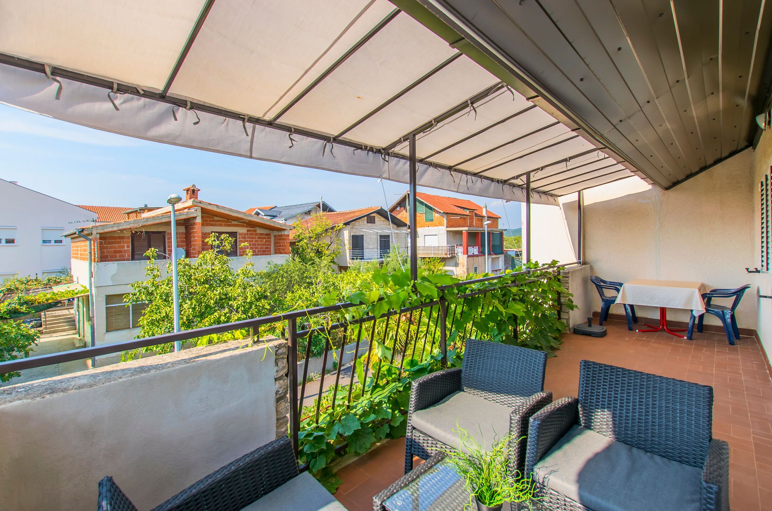 A covered balcony with wicker furniture, a table, and chairs, overlooking a residential area with houses and greenery.