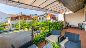 A covered balcony with wicker furniture, a table, and chairs, overlooking a residential area with houses and greenery. - Apartments Andrija (Pirovac)