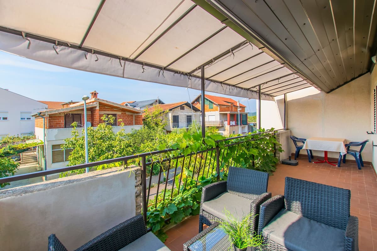 Ein überdachter Balkon mit Korbmöbeln, einem Tisch und Stühlen, mit Blick auf eine Wohngegend mit Häusern und Grünflächen.
