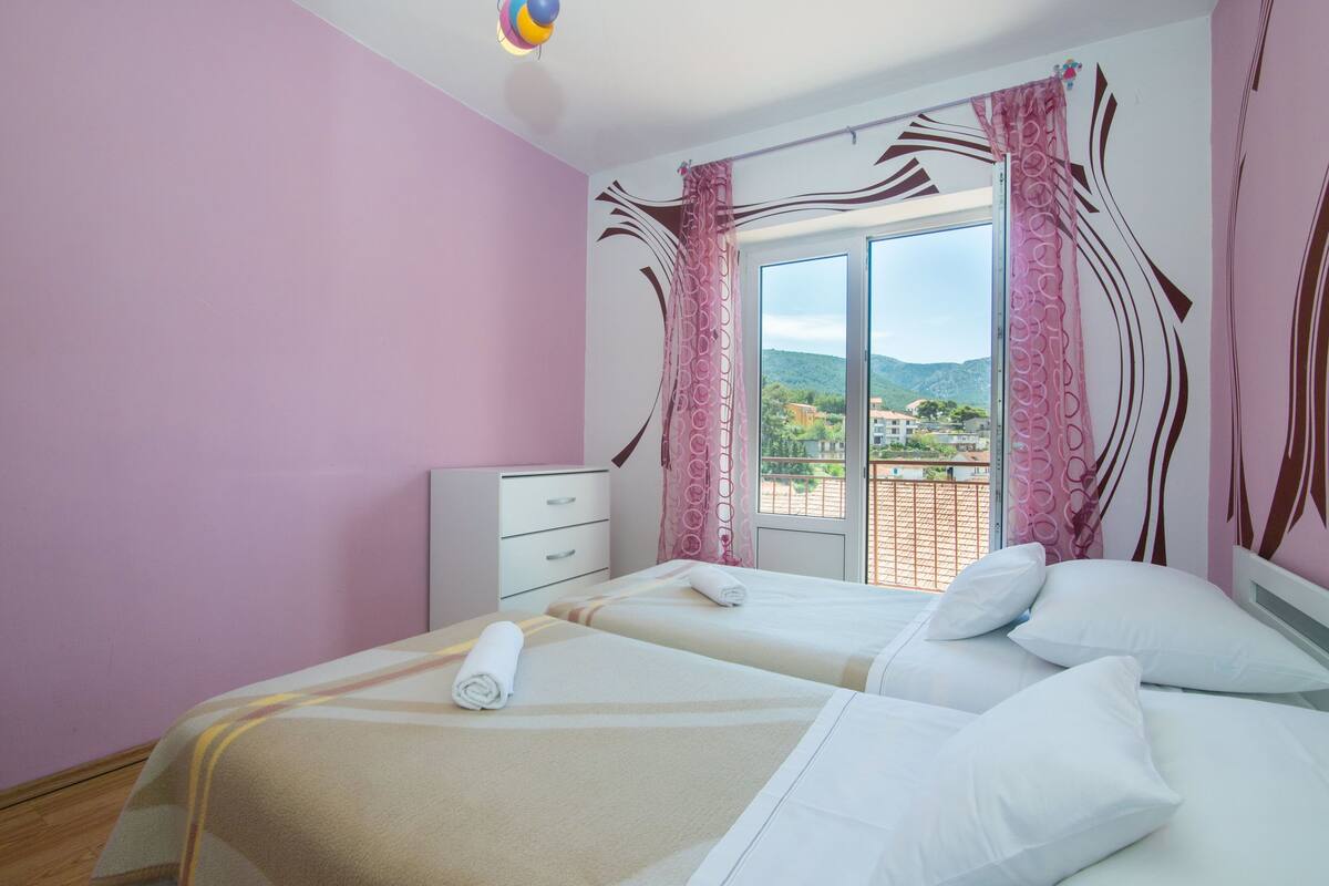 A hotel room with two beds, a window with curtains, a view of the city, and a balcony.