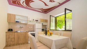 A compact kitchen with wooden cabinets, a dining table set for four, and a window with a view of greenery. - Apartments Jelka (Jelsa)
