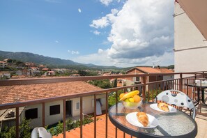 Un balcon avec une table dressée pour le petit-déjeuner, d’où l’on peut admirer une ville aux bâtiments aux toits rouges et des montagnes à l’horizon.