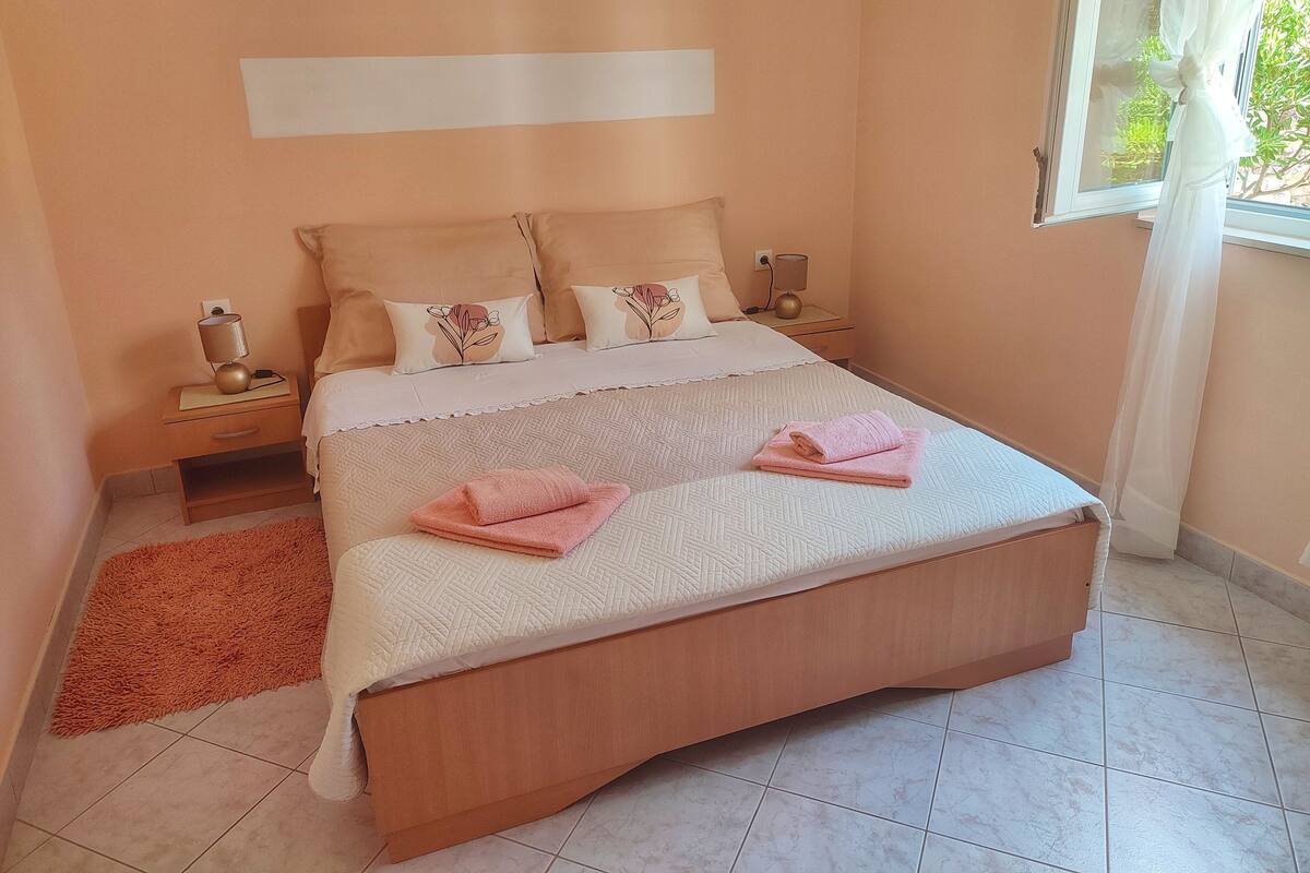 Una camera da letto con un letto grande, due comodini, un tavolino e una finestra con tende leggere, oltre a un piccolo tappeto arancione.