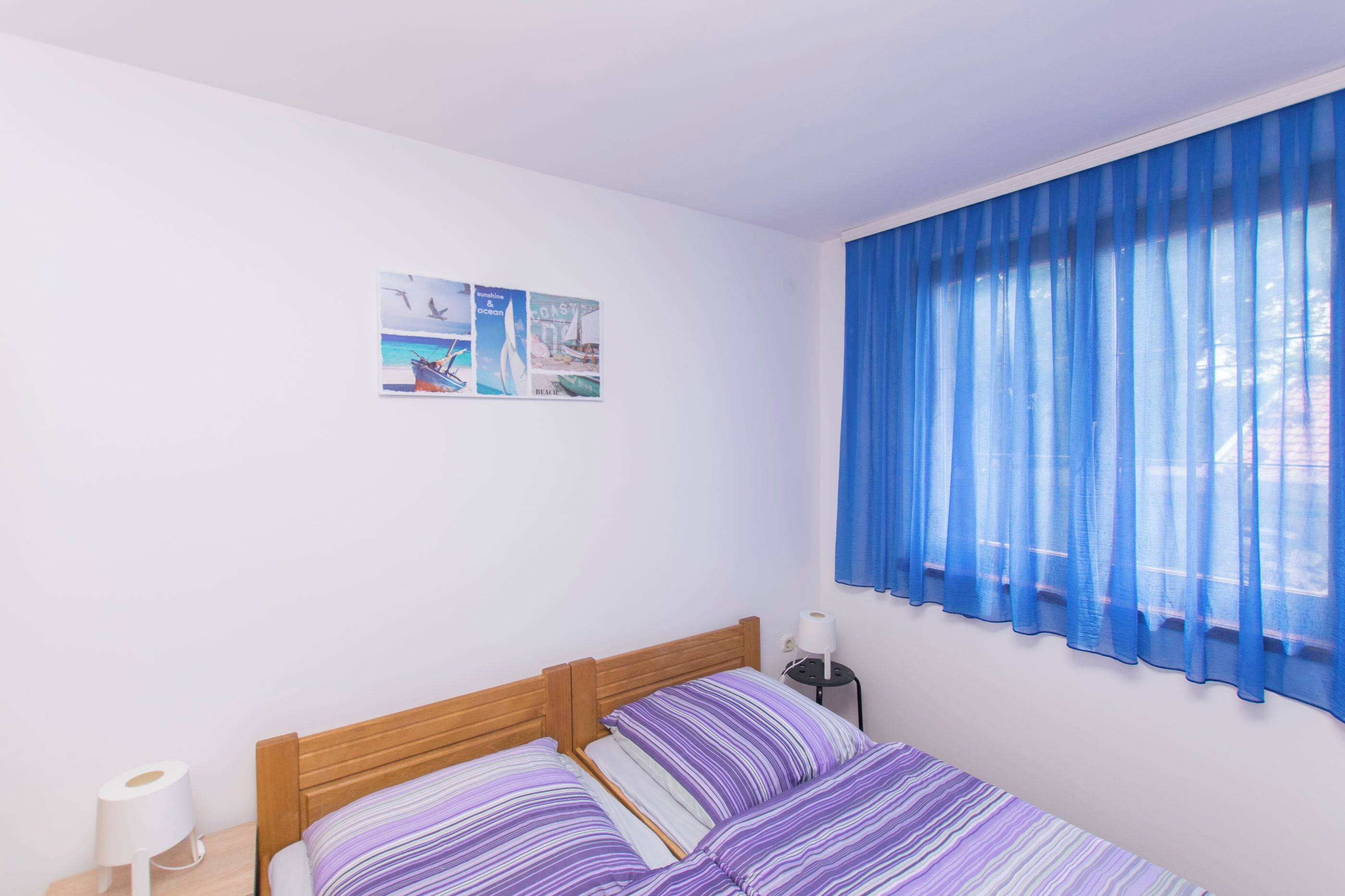 Ein Schlafzimmer mit einem hölzernen Bett, gestreifter Bettwäsche, einem Nachttisch mit Lampe und einem Fenster mit blauen Vorhängen.
