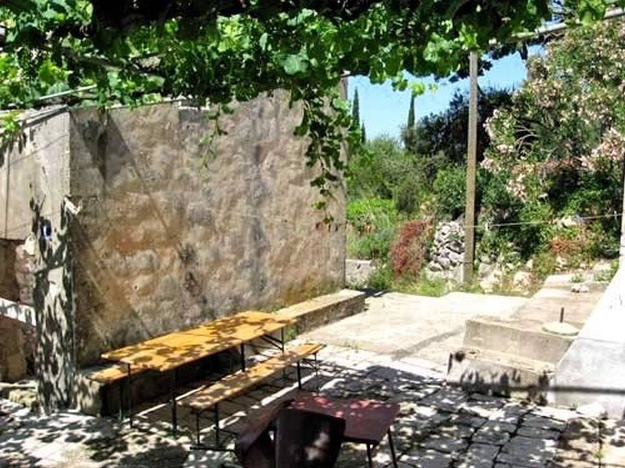 Un área de estar al aire libre con mesas y bancos de piedra, rodeada de vegetación y un muro de piedra.