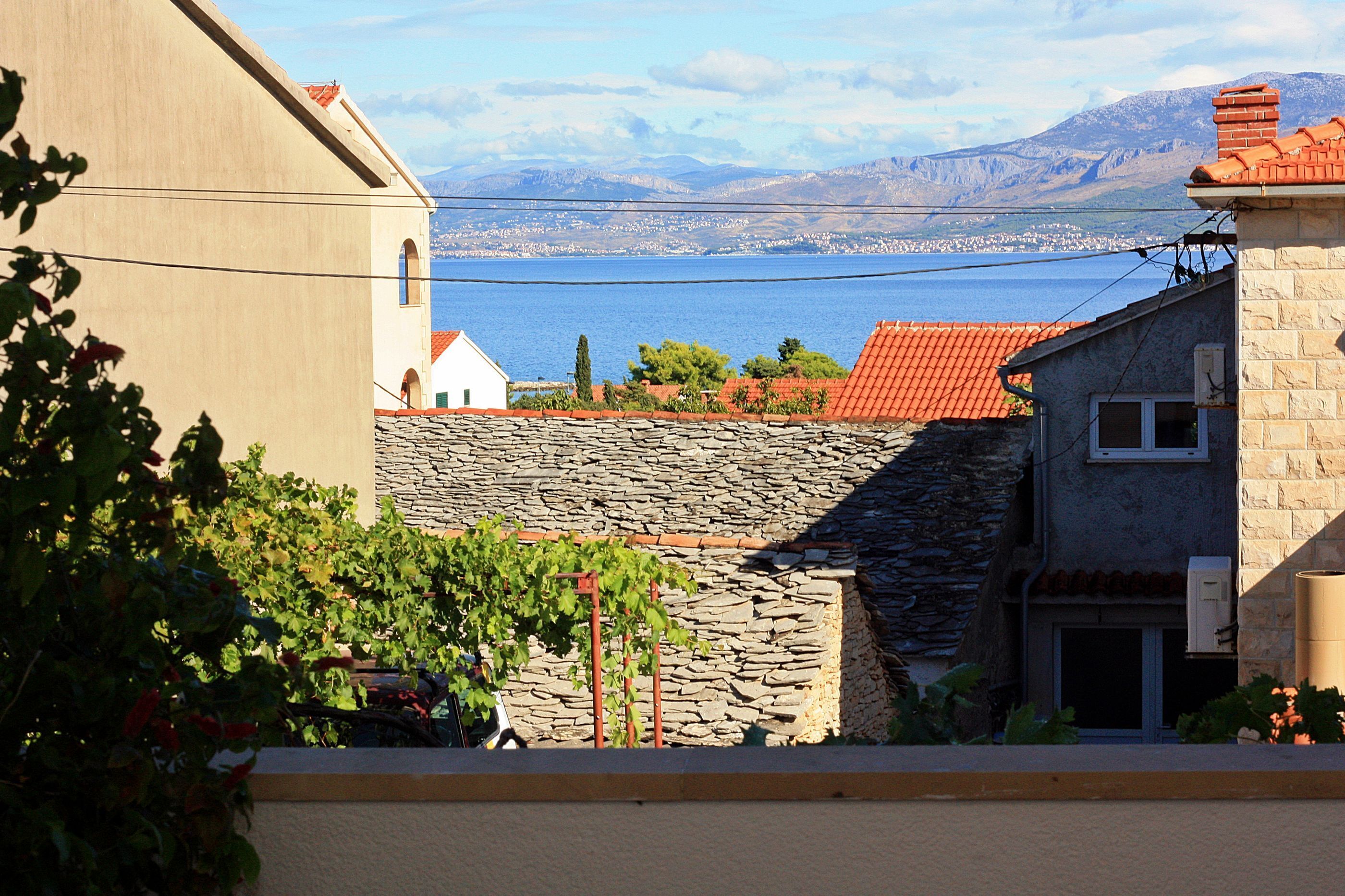 A coastal building with stone walls and a red-tiled roof, overlooking a body of water and distant mountains.