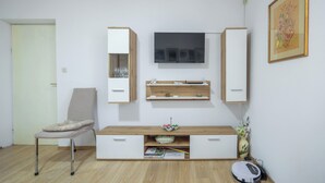 A modern living room with a flat-screen TV mounted on the wall, a wooden cabinet with shelves, a cushioned chair, and a small table with a vase and remote control. - Apartment Toma (Supetar)