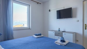 A bedroom with a large bed, a TV mounted on the wall, a window with curtains, and a small desk with a remote control. - Apartment Toma (Supetar)