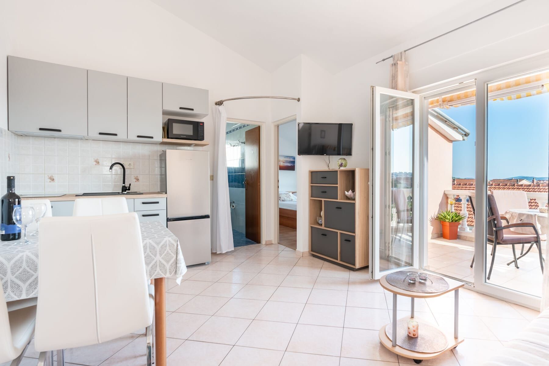 Una cocina moderna con armarios blancos, un microondas y un refrigerador. Hay un comedor con mesa y sillas, y un balcón con una mesa y sillas.