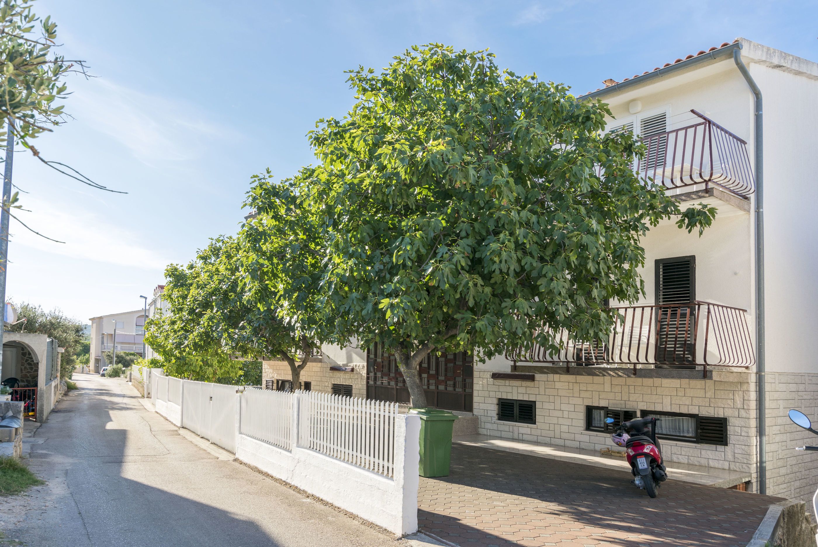 A narrow street lined with a white fence and a tree providing shade. A red scooter is parked on the sidewalk.