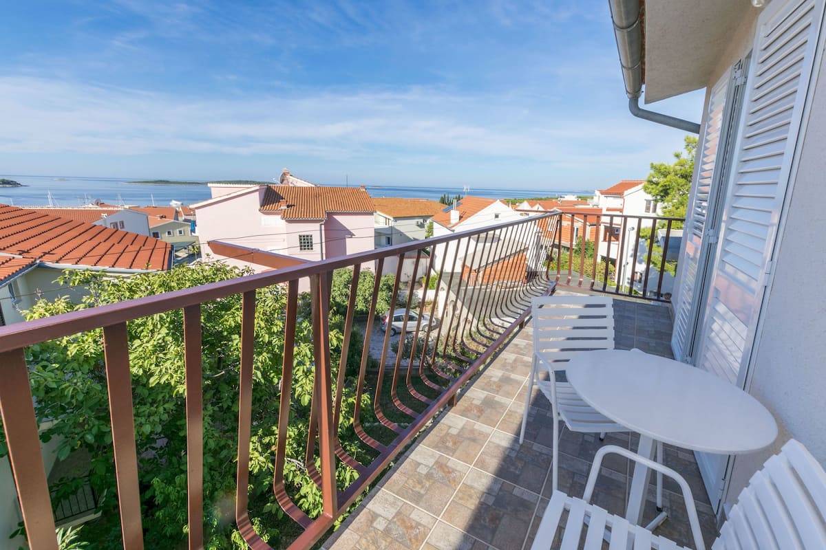 A balcony with a white table and chairs, overlooking a residential area with red-tiled roofs and a view of the sea.