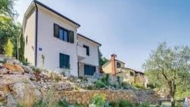 A modern two-story house with a stone retaining wall, surrounded by greenery and trees. - Apartments Grzic (Porozina)