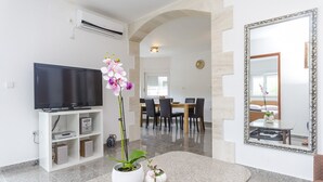 Una sala de estar moderna con un televisor de pantalla plana, un mueble blanco para televisión, una zona de comedor con una mesa y sillas, y un espejo grande en la pared.