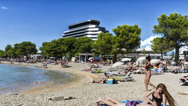 Una playa de guijarros con gente tomando el sol, sombrillas y un edificio moderno al fondo.
