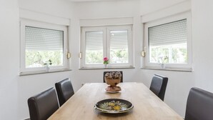 Comedor con mesa de madera, sillas negras y un cuenco decorativo. Hay tres ventanas con persianas, cada una con una maceta en el alféizar.