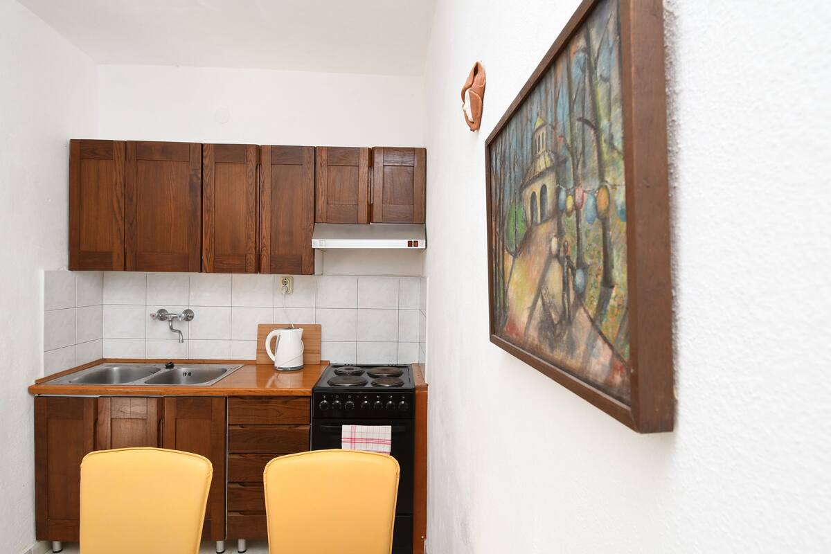A compact kitchen with wooden cabinets, a stove, two yellow chairs, and a painting on the wall.