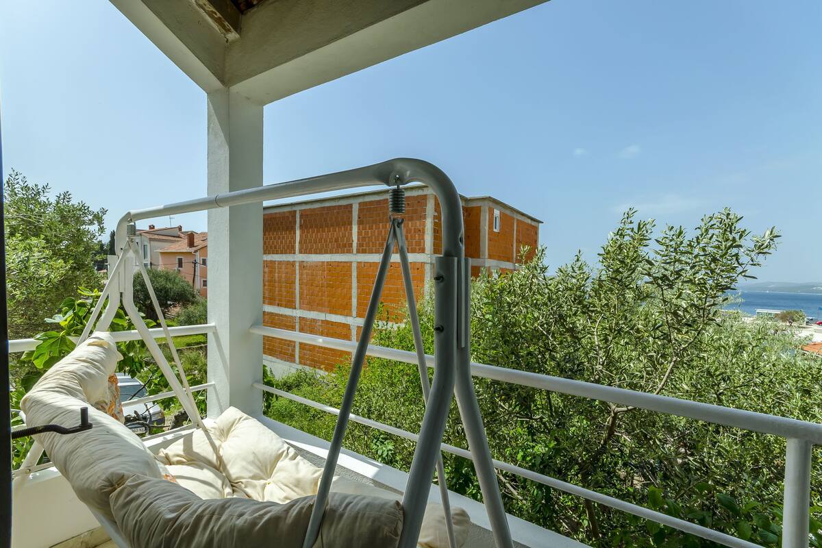 A balcony with a metal railing, a cushioned bench, and a view of a brick building and greenery.