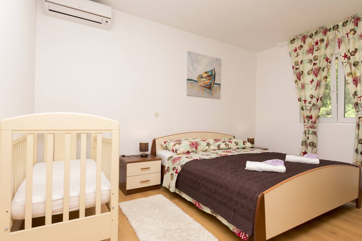 A bedroom with a crib, a bed with a floral bedspread, a painting on the wall, and a window with curtains.