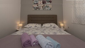 Ein ordentlich bezogenes Bett mit floralem Bettwäschemuster, zwei Nachttischlampen und drei gerollte Handtücher in Lila, Blau und Hellblau.