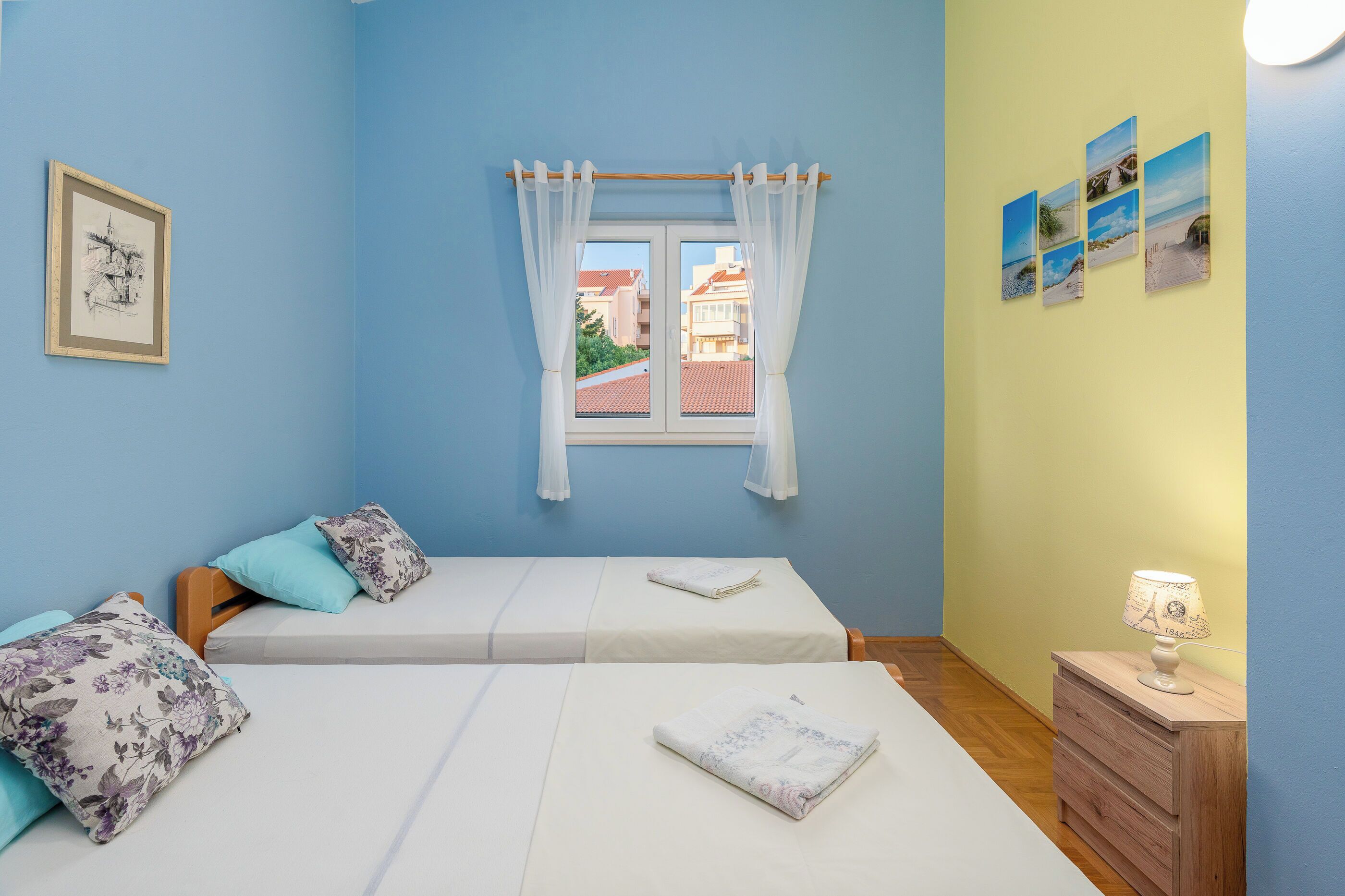 Una habitación con dos camas, una ventana con cortinas, una pared con cuadros y una mesita de noche con una lámpara.