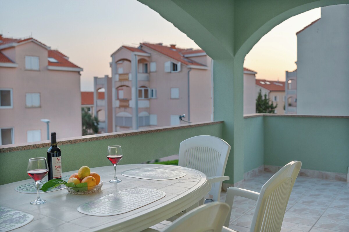 A balcony with a table set for two, featuring a bottle of wine, glasses, and a fruit bowl.