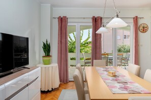 A modern kitchen with a flat-screen TV, a dining table with chairs, a potted plant, and a view of the outdoors through large windows. - Apartments Jasna (Novalja)