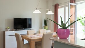 Ett modernt matområde med ett träbord och stolar, en rosa krukväxt och en platt-TV placerad på ett vitt skåp.