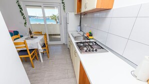 A compact kitchen with a gas stove, a sink, and a dining area with a table and chairs. - Apartments Jerkunica (Rogoznica)