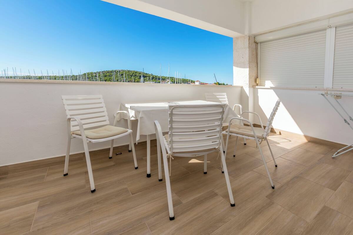 Ein Balkon mit weißen Stühlen und einem Tisch, Blick auf das Meer sowie eine geschlossene Rolltür.