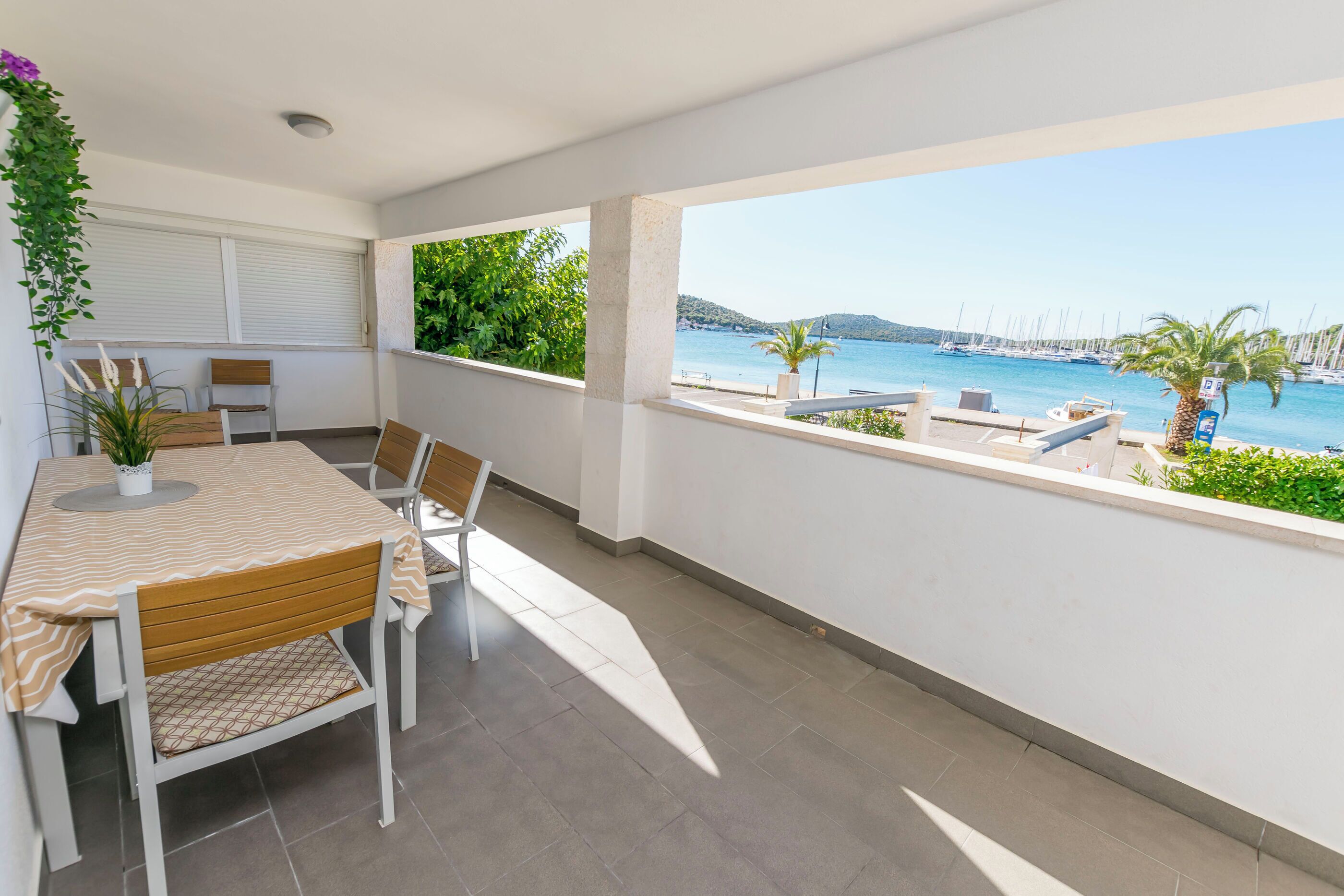 Un balcone con tavolo e sedie, affacciato su una marina con barche e cielo sereno.