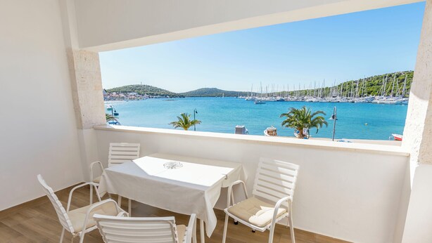A balcony with a table and chairs, overlooking a marina with boats and a hilly landscape. - Apartments Jerkunica (Rogoznica)