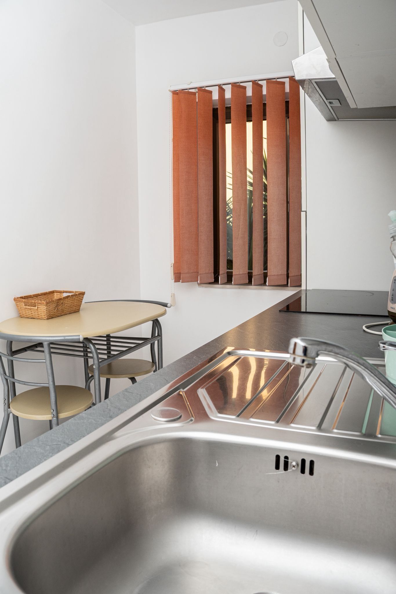 A modern kitchen with a stainless steel sink, a small round table with a basket, and a window with vertical blinds.