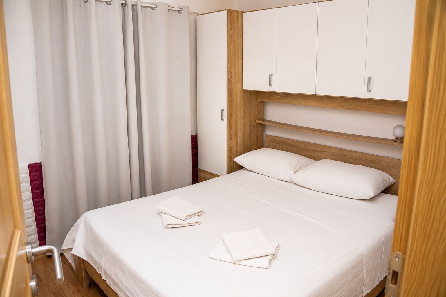 A neatly made bed with white linens and two folded towels, a wooden headboard, and a white wardrobe with silver handles.